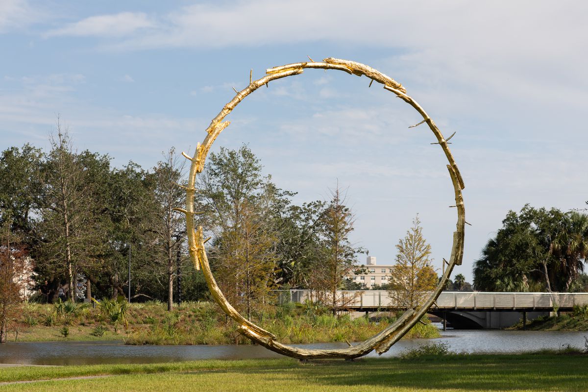 A large gold circular sculpture in a park with trees and lagoons