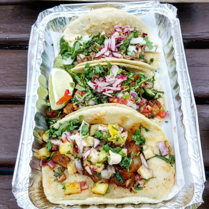 Three tacos in an aluminum takeout container on a picnic table.