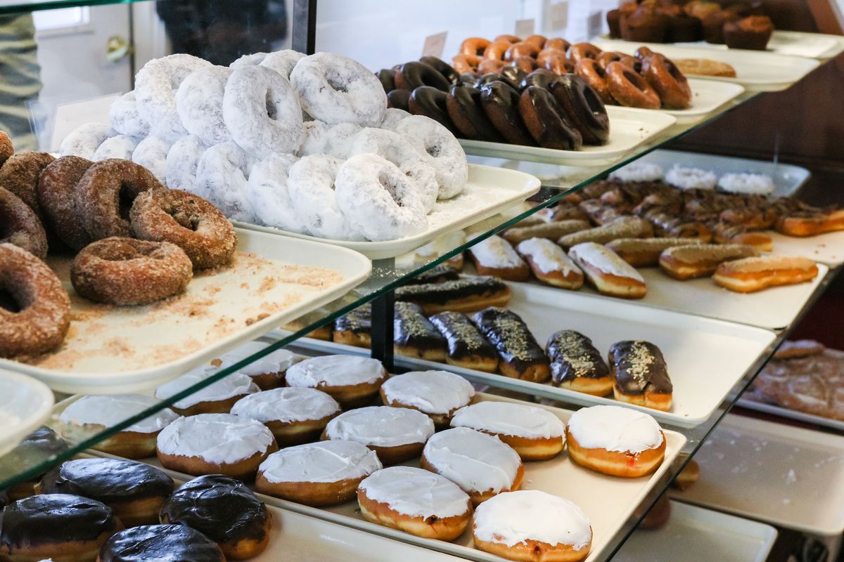 Two shelves stacked with trays of various cake and glazed doughnuts.