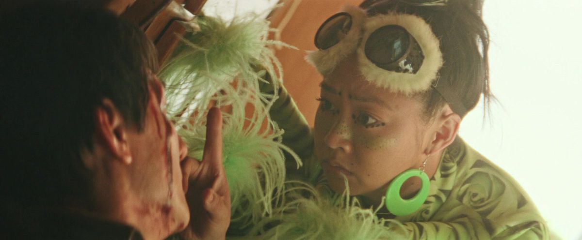 Jobu Tupaki aka Joy Wang (Stephanie Hsu) wearing furry eye goggles pulled up her forehead, big green ring earrings and a green frill shirt holds a finger up to Waymond (Ke Huy Quan) who is bleeding on the side of his face in Everything Everywhere All at Once