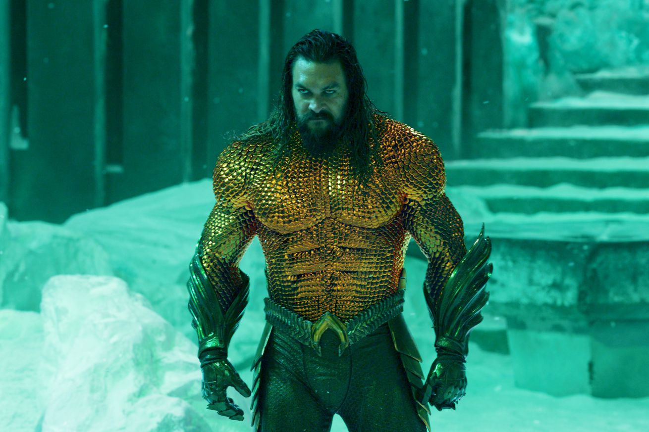 A man with long, scraggly brown hair wearing a superhero costume consisting of skin-tight orange armor made of what appears to be large orange scales across his upper body, and large green scales down his lower arms and legs. The man is standing in a frozen wasteland.