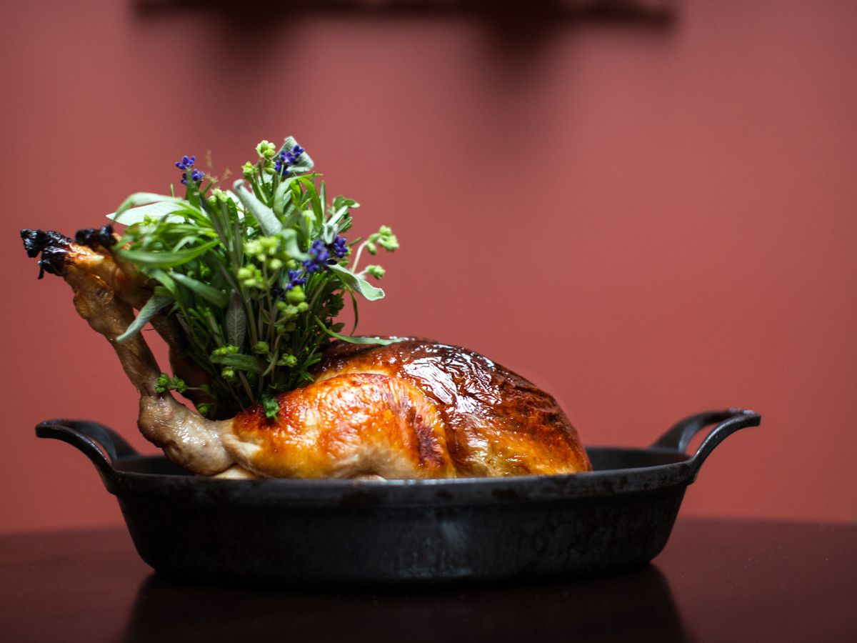 A whole roast chicken in a black roasting pan in front of a red background.