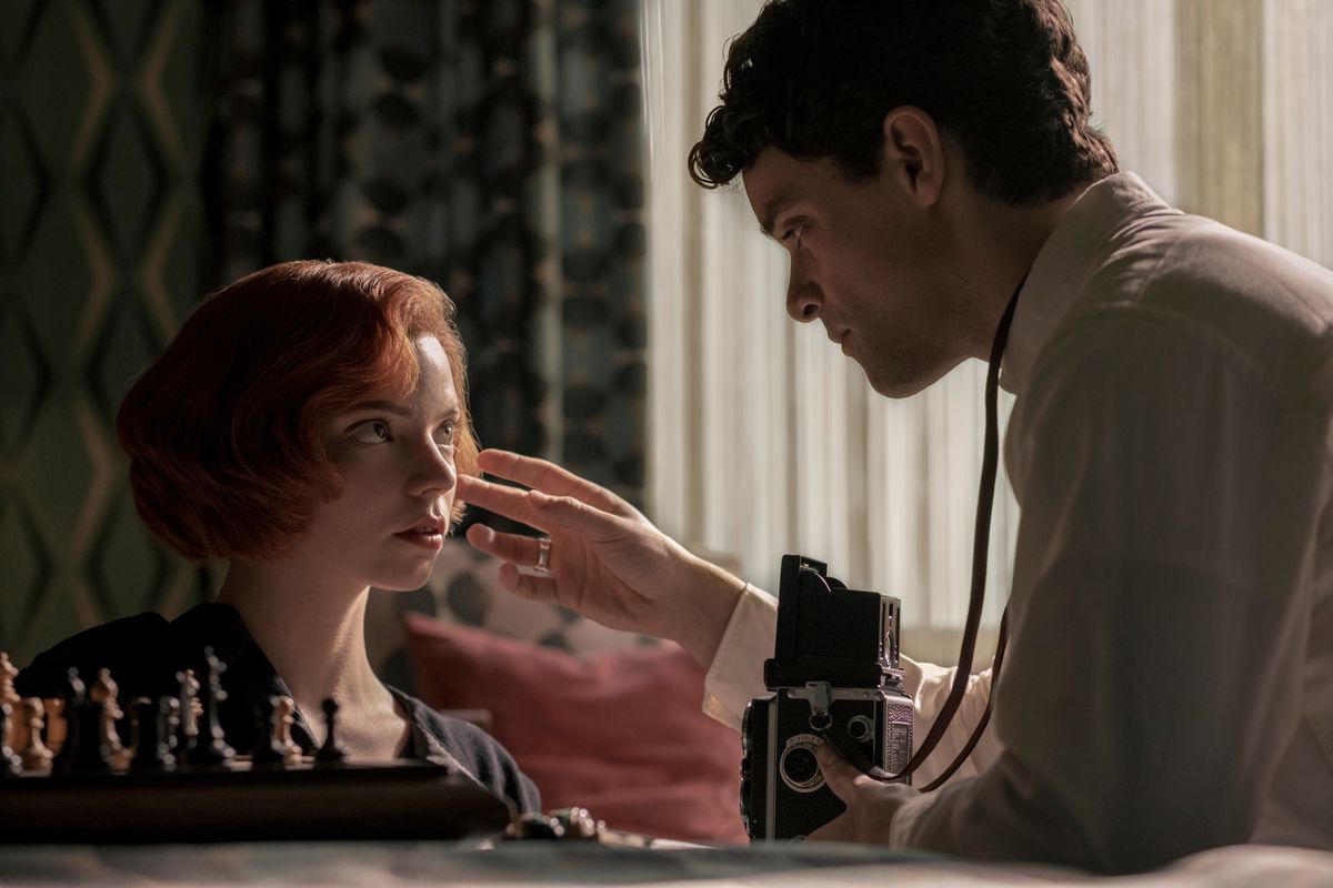 A red-headed woman (Beth) kneels before a chess board perched on the side of a bed. A brown-haired man holding a camera adjusts her hair as he prepares to photograph her.