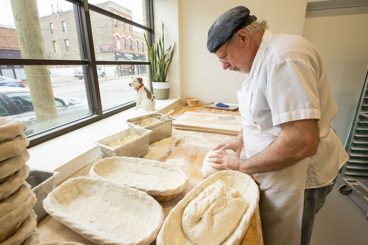 A baker stands at a countertop shaping dough into an oblong loaf and prepares to put it into a basket.