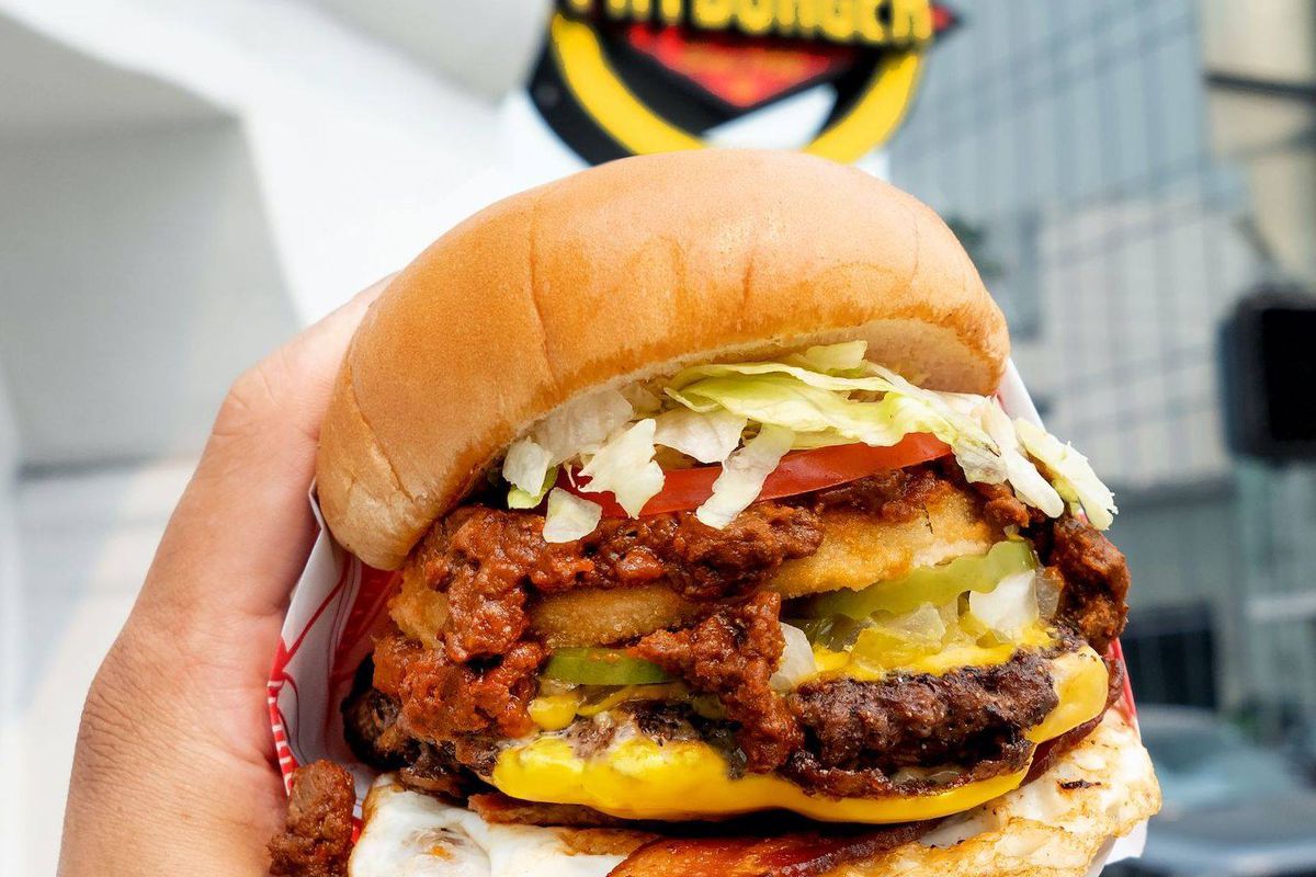 a hand holding a burger topped with chili and a fried egg in front of a fatburger neon sign
