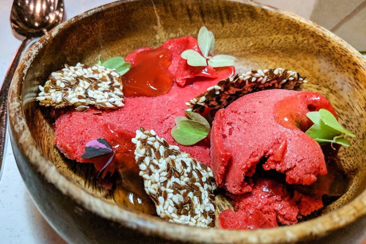 Several scoops of bright pink raspberry sorbet are served in a wooden bowl and garnished with seeded crackers and edible flowers.