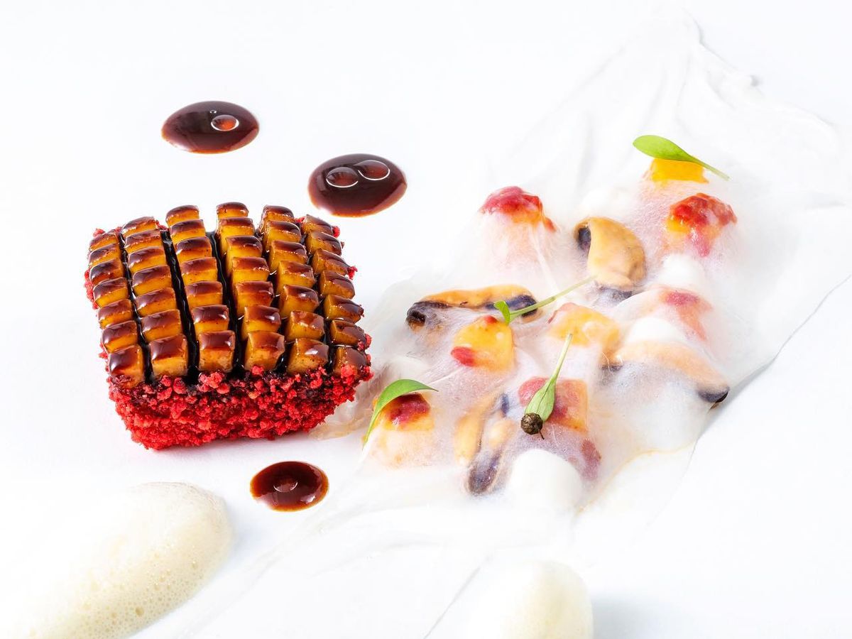 Geometrically sliced squid with a bright red crust sits beside chunks of more squid, flowers, and mango slices beneath a white foam.