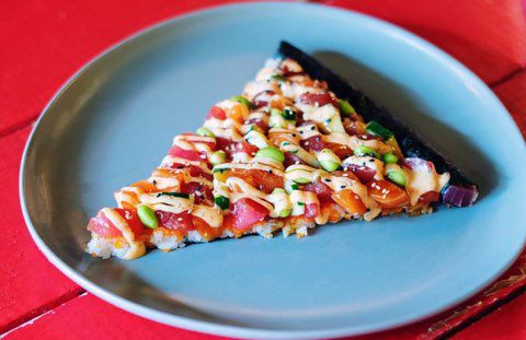 A slice of sushi pizza on a blue plate sits on a bright red table. 