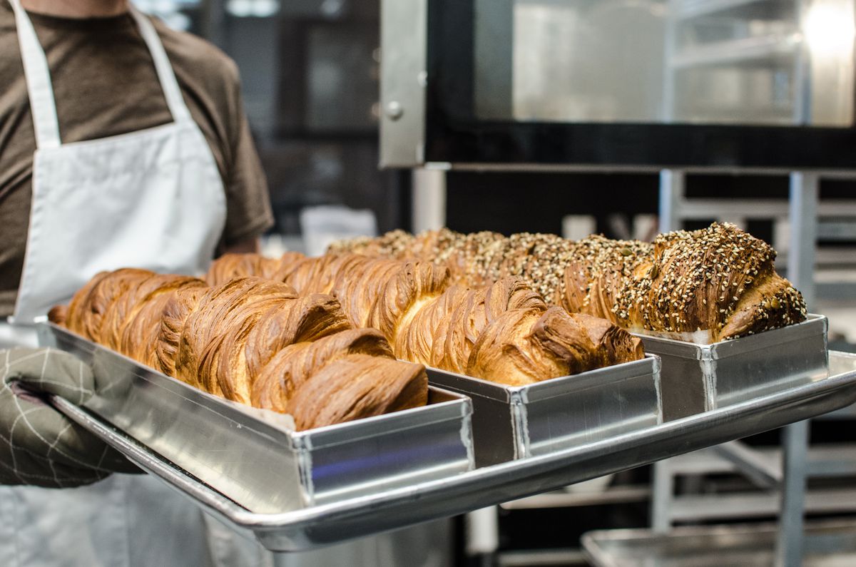 Visible from the neck down, a person in a white apron and heavy oven mitts holds a metal tray of three loaves of babka just outside an open oven door.
