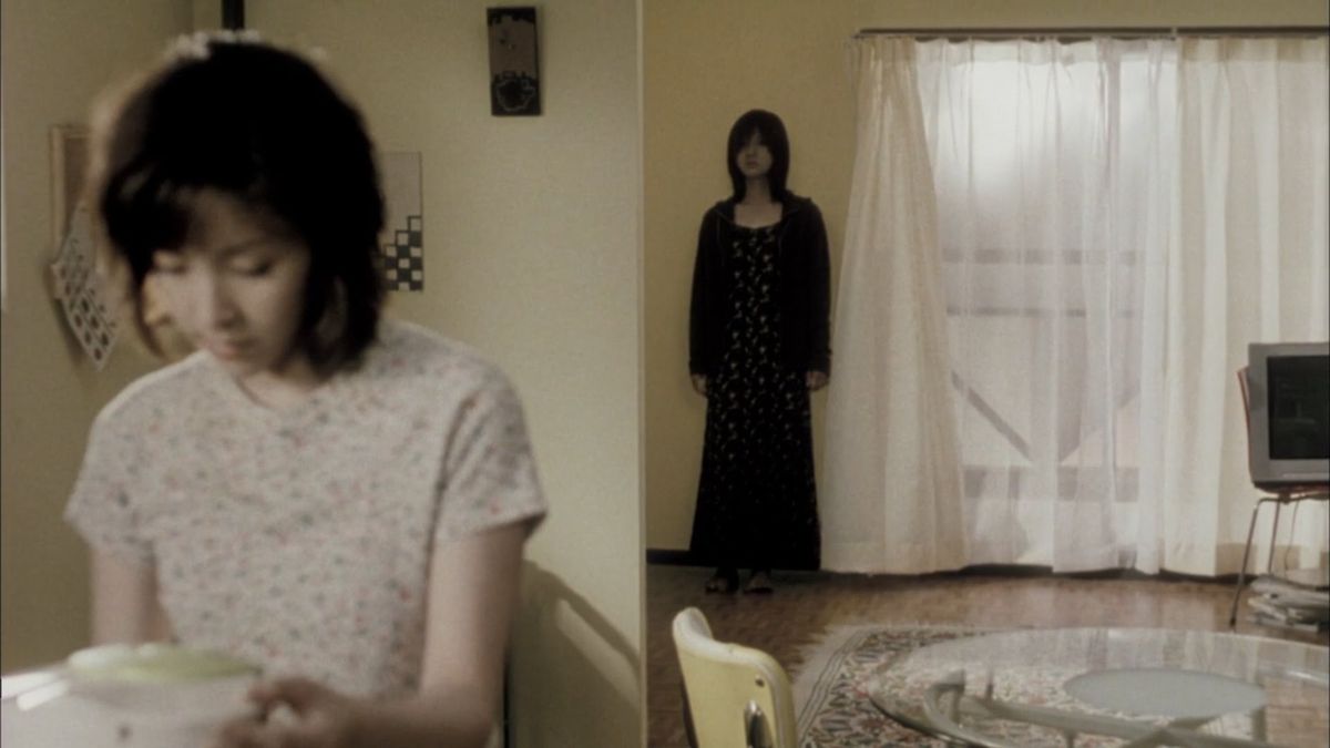 Eine Frau in Schwarz steht bedrohlich hinter einer ahnungslosen Frau in ihrer Wohnung in Pulse.