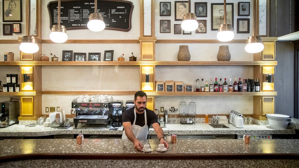 A barista sets down two small cups of coffee underneath overhead light fixtures, vintage photos, and a sandwich board menu