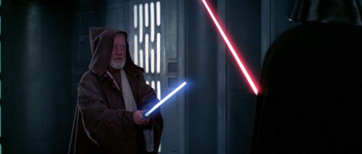 1977 Obi-Wan Kenobi (Alec Guinness), in Jedi robes and with his pointy hood pulled up to partially conceal his face, holds up his lightsaber as Darth Vader looms at the edge of the image in Star Wars: A New Hope