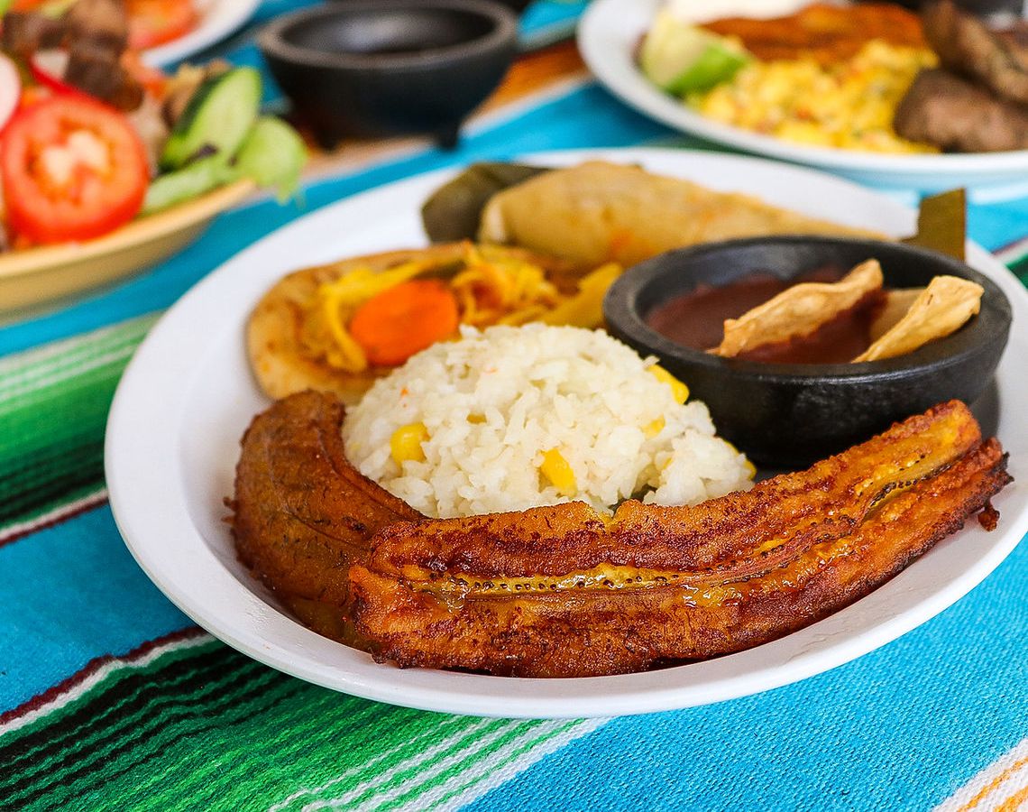 A plate for rice, vegetables, salsa, and a long plantain on a bright striped table cloth. 
