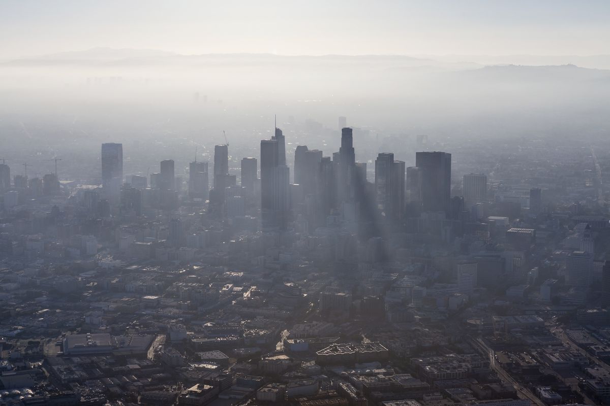 An aerial view of the cityscape of Los Angeles. There is a heavy layer of smog hanging over the buildings.
