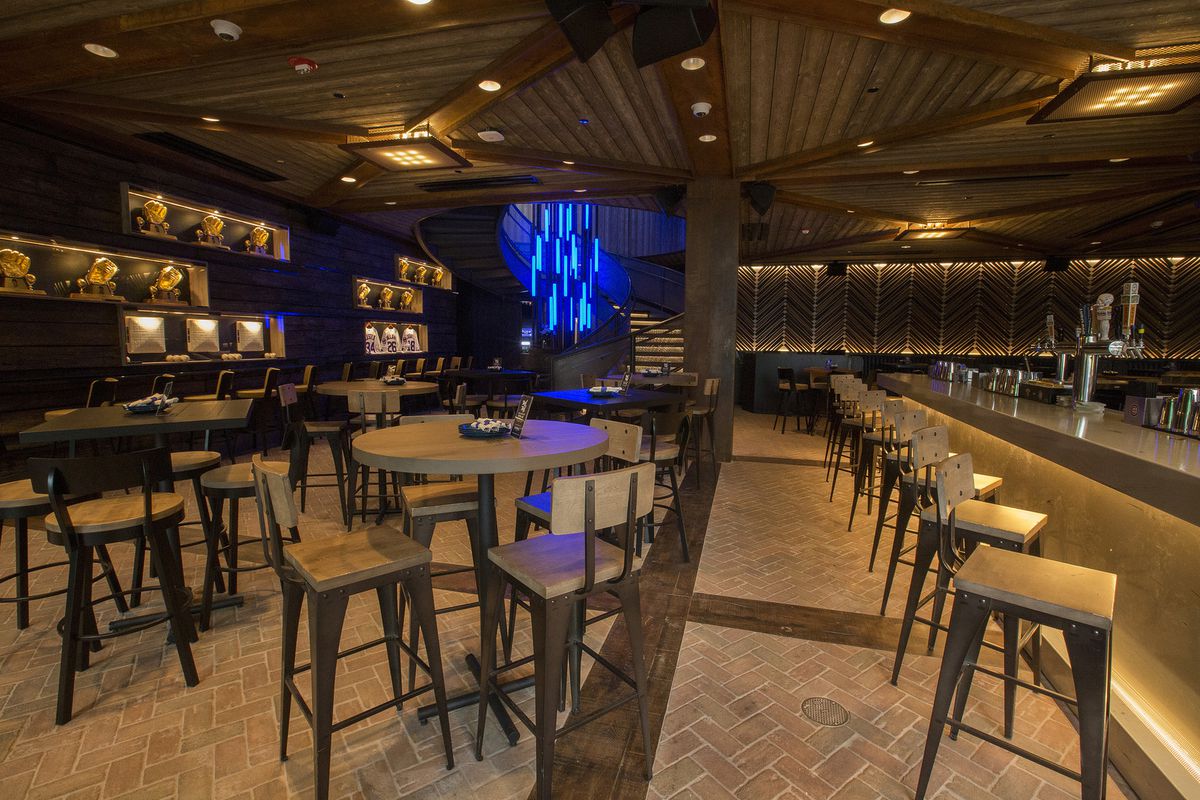 A bar space with exposed brick flooring, high-top tables and chairs, and warm yellow lighting. The space has a wood paneled ceiling and a glowing blue light fixture sits in a back corner.