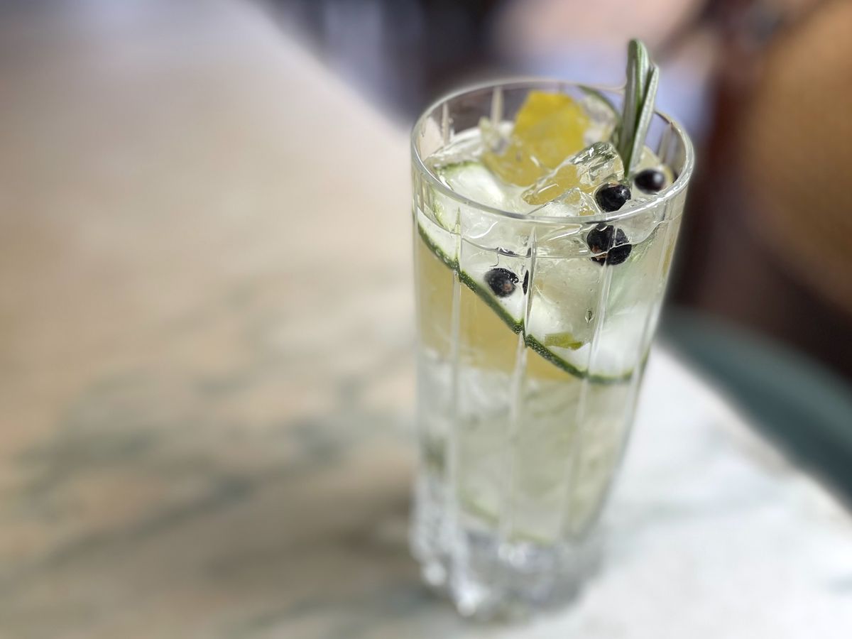 A highball glass is filled with a clear liquid, garnished with a cucumber ribbon, lemon zest and a few juniper berries. The glass sits on a marble topped table. The historic wood filled interior of Frost’s bar is barely visible in the background.