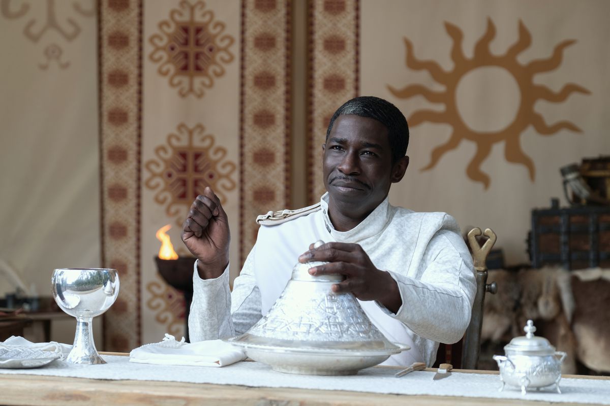 Abdul Salis as Whitecloak Questioner Eamon Valda in The Wheel of Time season 1, sitting down at a table and reaching for a plate