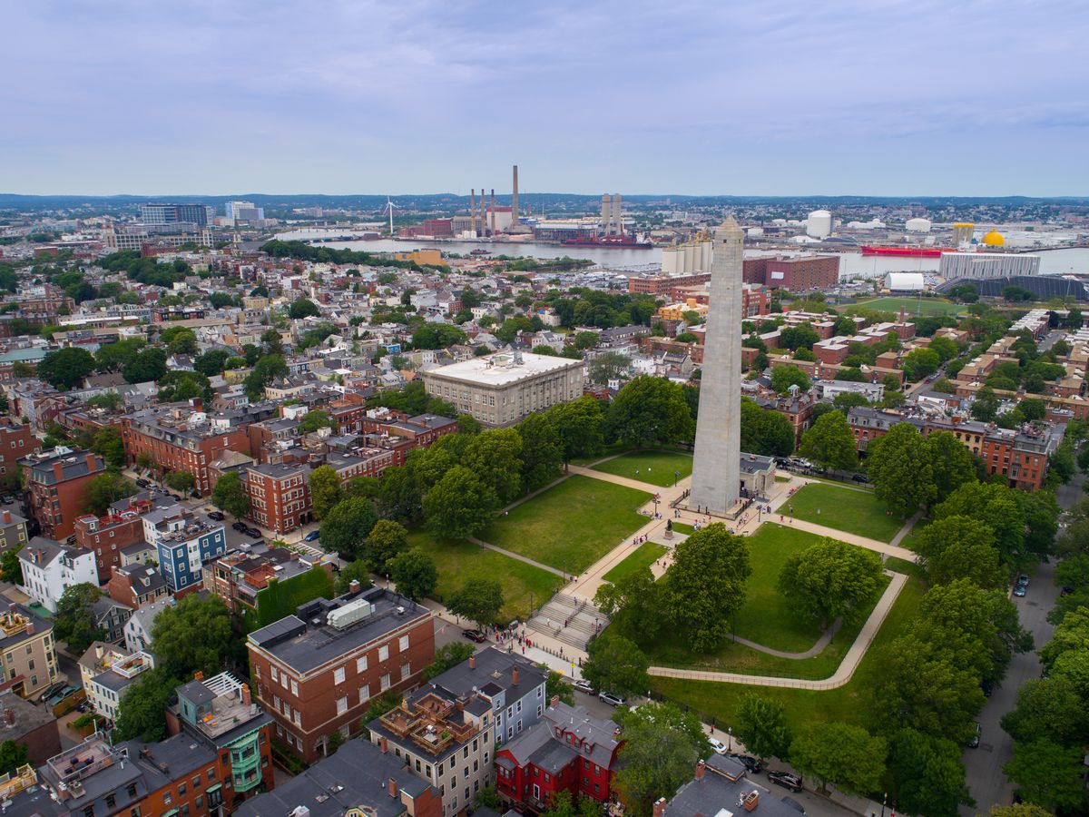 An aerial view of a large monument surrounded by park space. The park space is surrounded by various city buildings in Boston.
