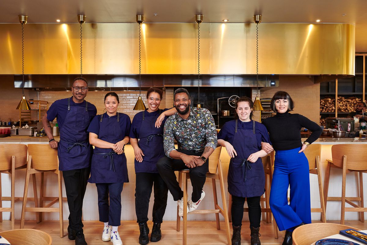 A smiling, diverse kitchen team stands in front of the kitchen, wearing blue aprons and surrounding the chef, who wears a floral shirt.