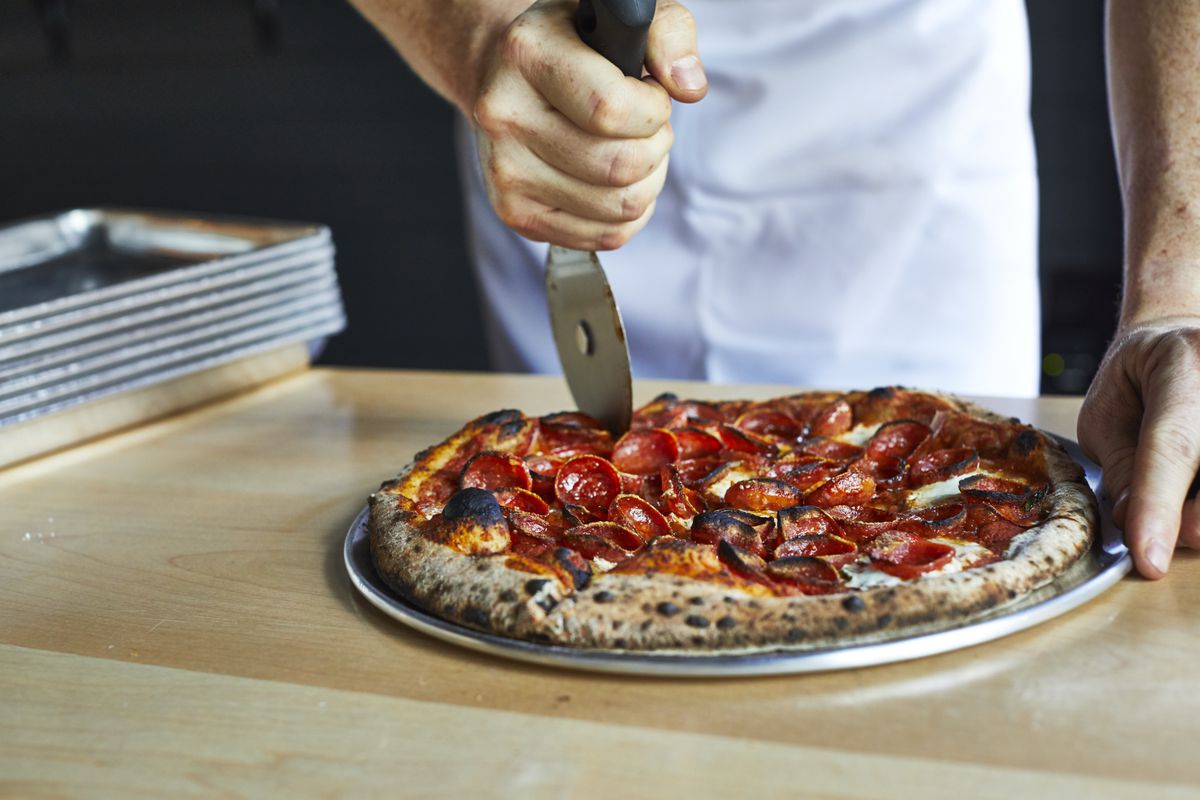a man’s hand slices a pepperoni topped pizza on the counter