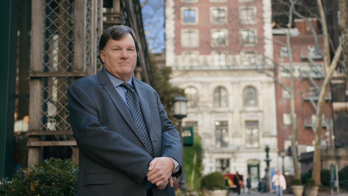 Rex Heuermann in a gray suit poses for a photo in front of a brick Manhattan building.