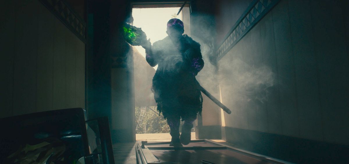 Peter Dinklage as Toxie in The Toxic Avenger, shrouded in shadow and holding a crystal green staff while walking through a doorway