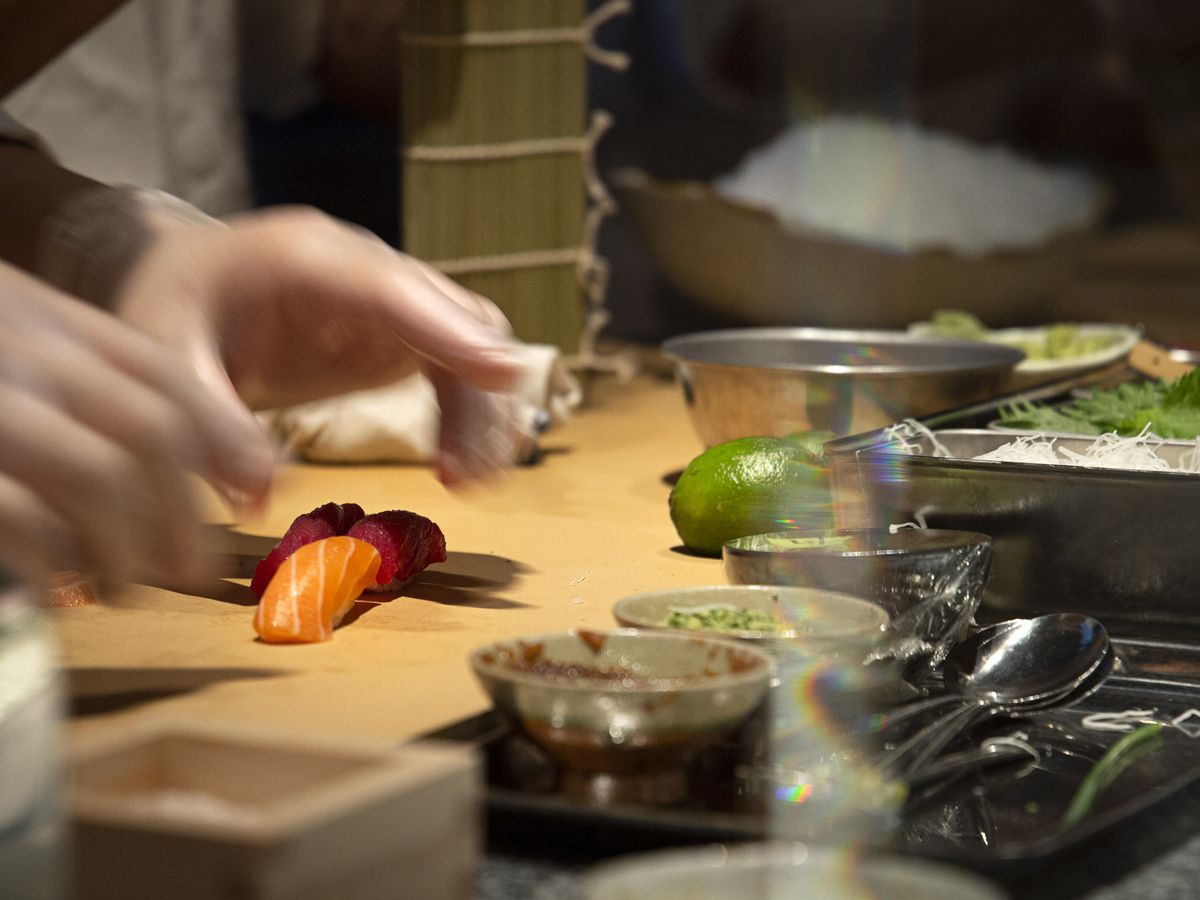 Hands reach towards a piece of sushi set on a cutting board surrounded by dishes of indiscernible ingredients