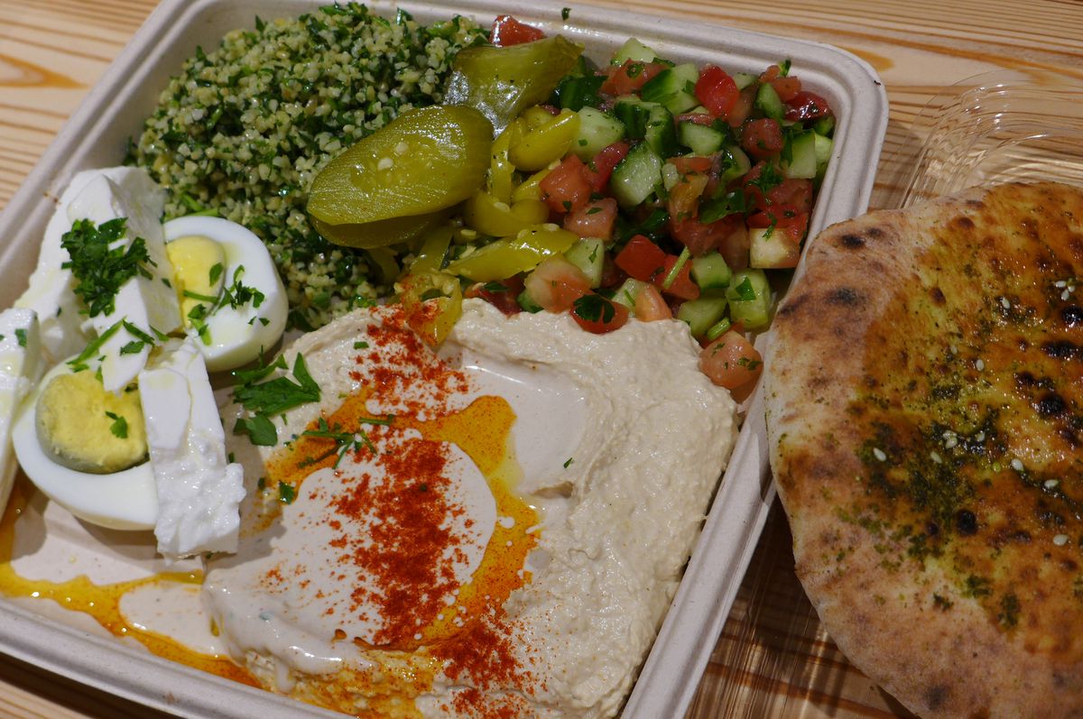 A plate with hummus with red powder and oil sprinkled on top, a cut up boiled egg next to it, along with greens and a piece of pita.