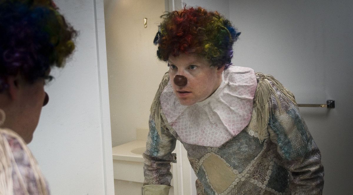 A man in a dark clown suit looks into the mirror in alarm in Clown