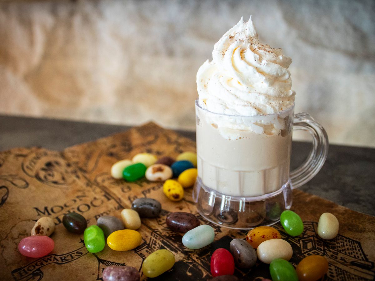 A mug of butter beer topped with a swirl of whipped cream surrounded by various jelly beans.
