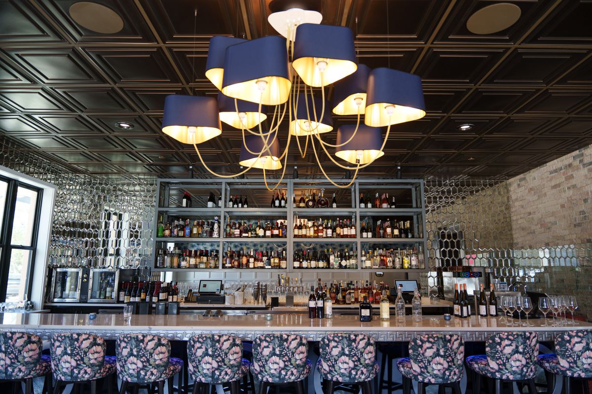 A long bar with floral fabric chairs and a chandelier in the foreground. 
