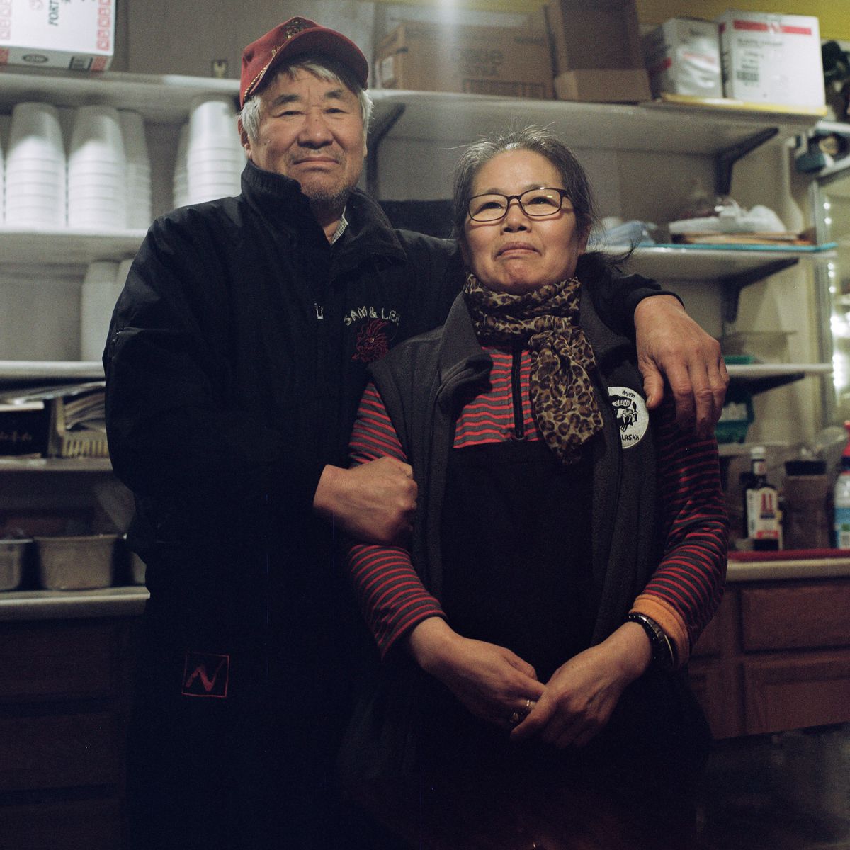 Older Korean man in a red cap stands and smiles with his arms around an older korean woman in front of commercial restaurant kitchen shelves.