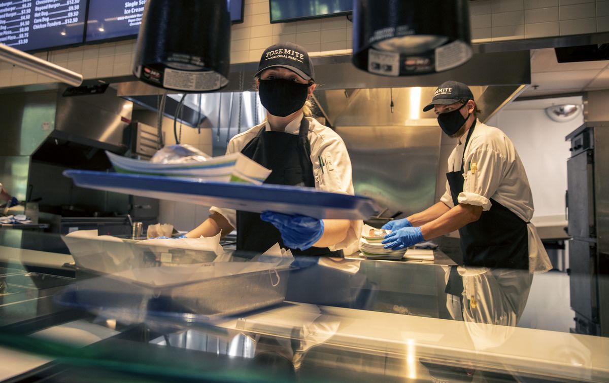 Woman standing behind restaurant counter wearing a mask and cap that reads “Yosemite.”