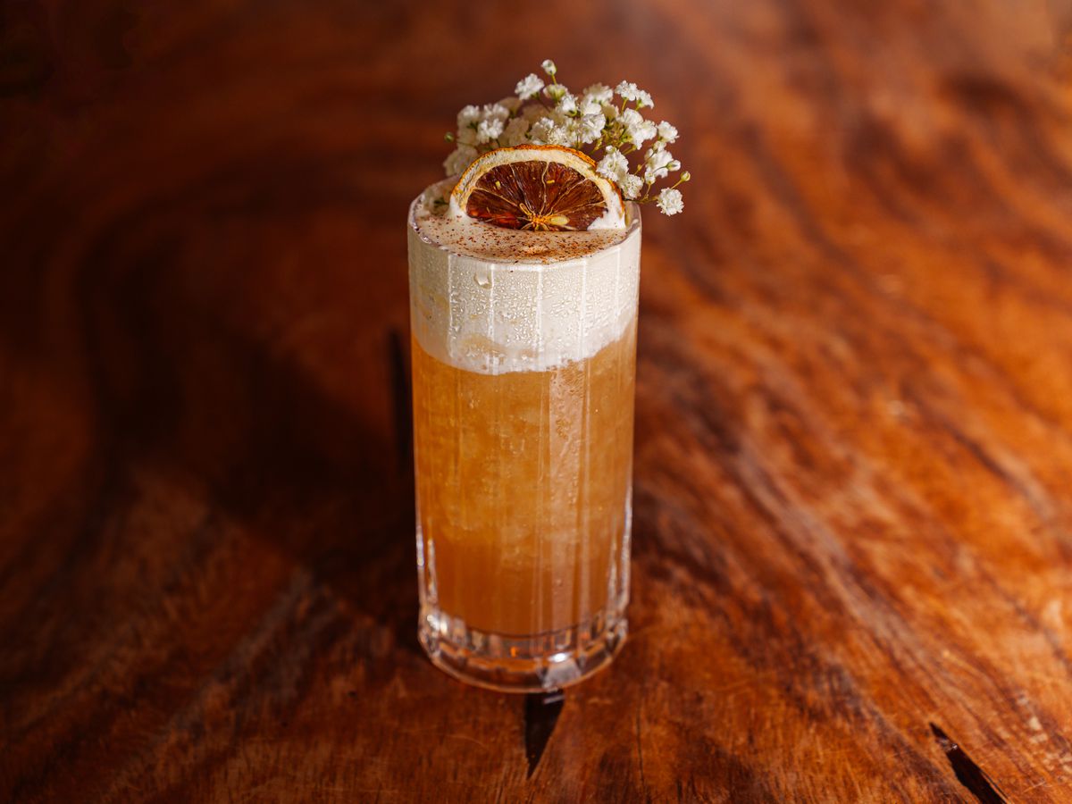An iced cocktail in a highball glass, topped with foam, an orange slice, and stems of baby’s breath.