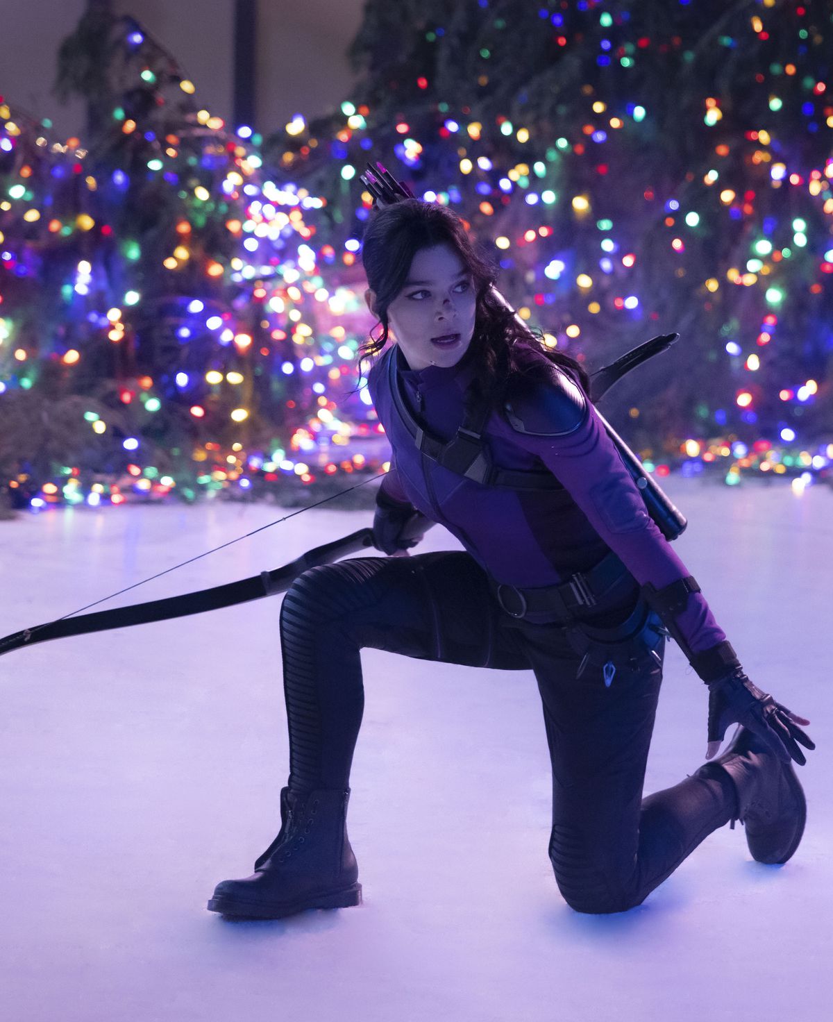 Kate Bishop (Hailee Steinfeld) in her purple uniform kneels on the ice of an ice skating rink while holding her bow, while christmas lights illuminate her from behind