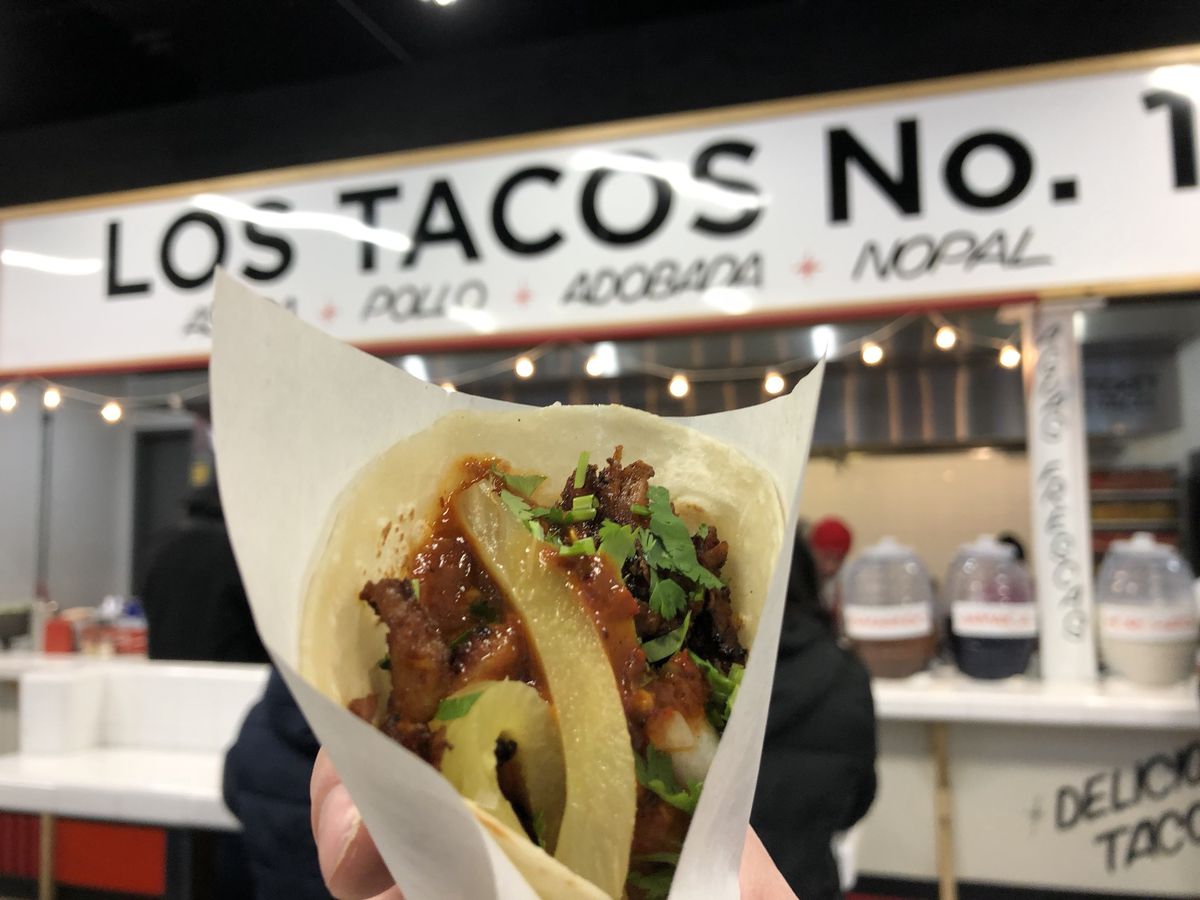 An adobada taco in a flour tortilla held up in the foreground with the Los Tacos No. 1 restaurant sign in the background