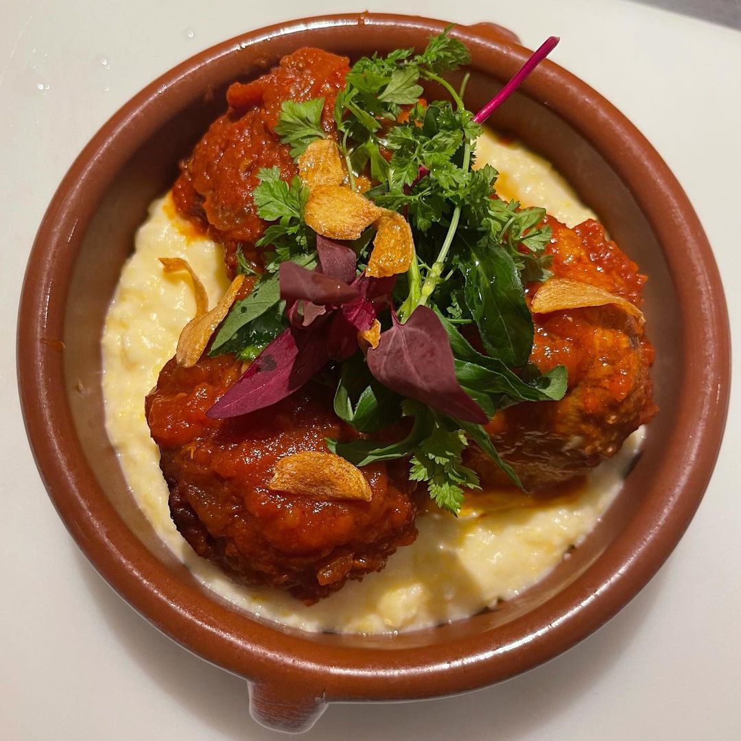 Three meatballs in tomato sauce are nestled on a bed of cheesy polenta in a small round brown bowl.