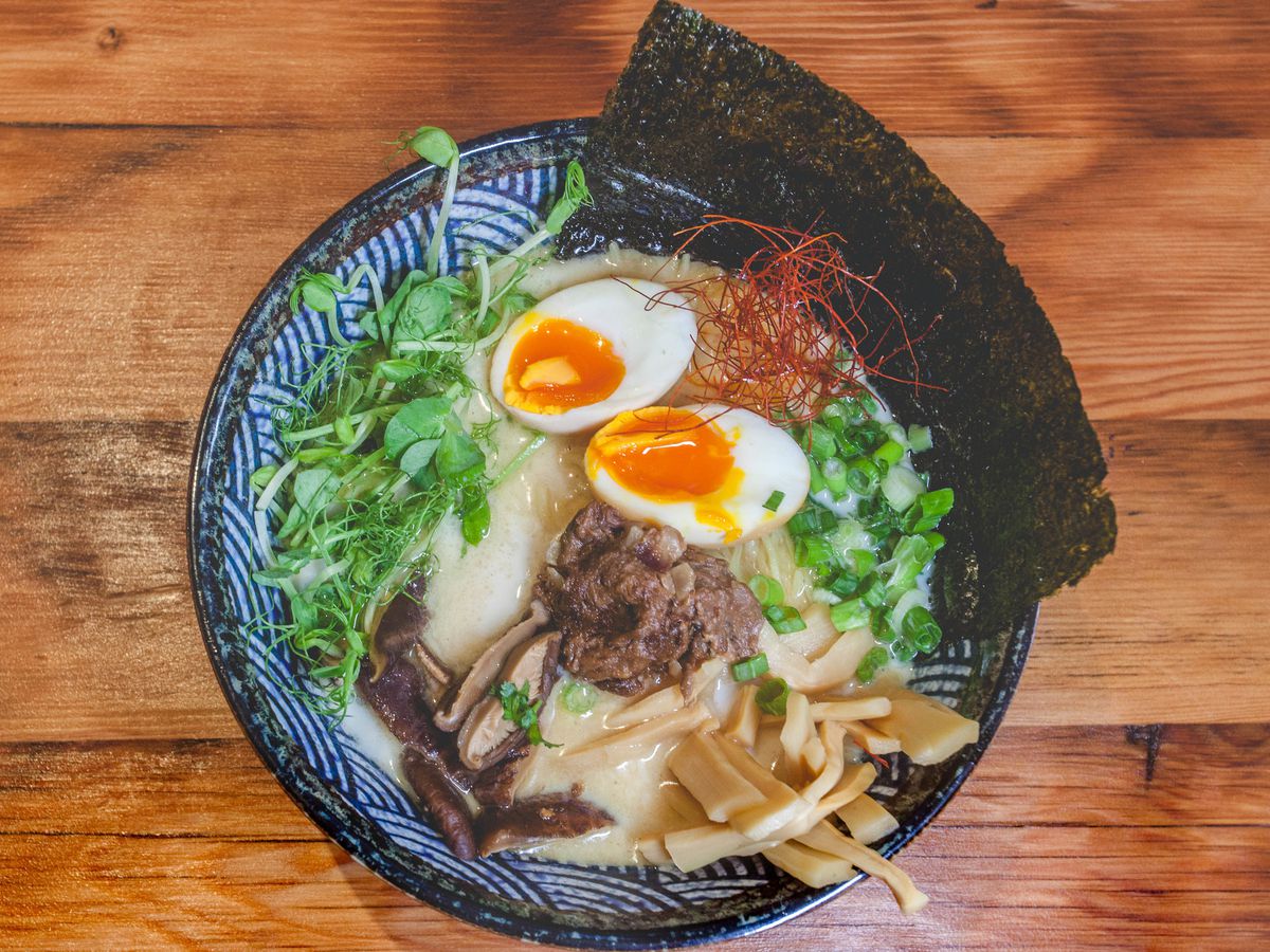A bowl of oxtail kare kare ramen shot from above, with nori, soft-boiled eggs, pea shoots, spring onions, and shiitake mushrooms