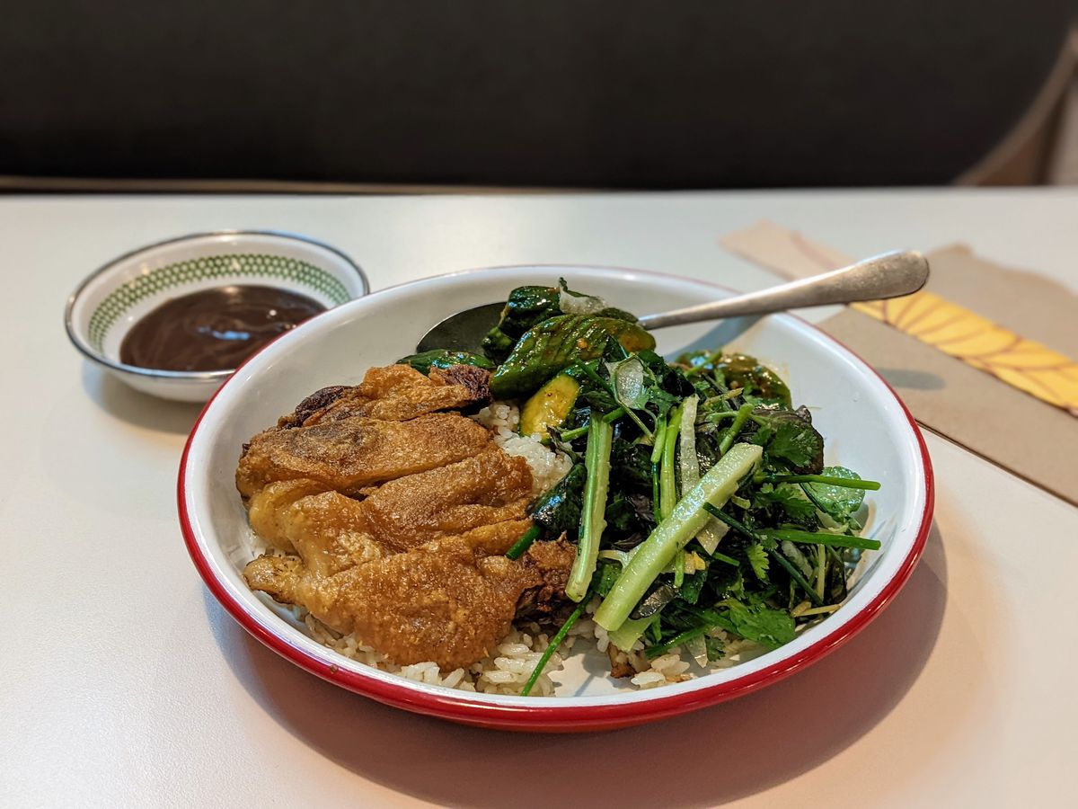 A white bowl with a crispy duck leg over rice with green vegetables and a side of hoisin sauce.
