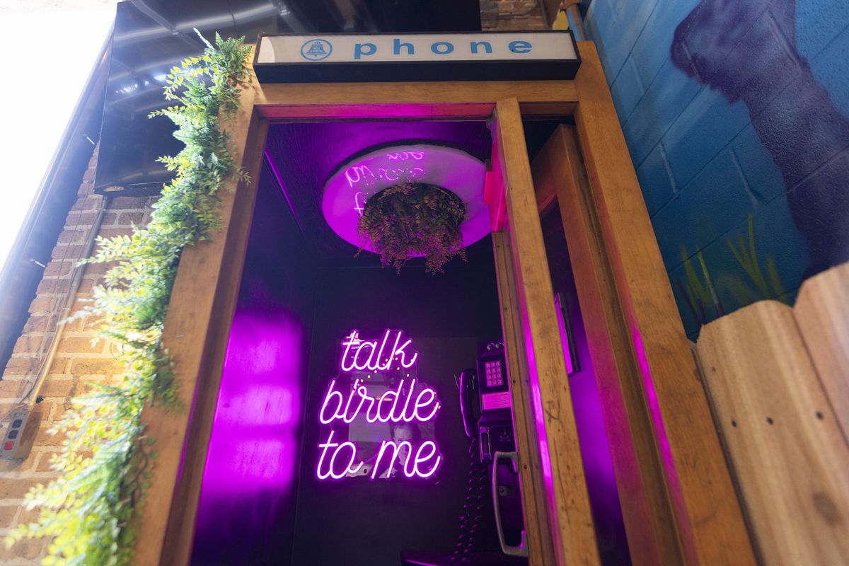 A wooden phone booth with a pink neon sign that reads “Talk birdie to me.”