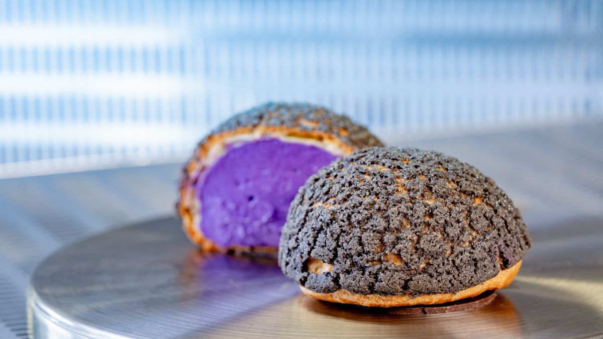 A large cream puff filled with purple cream and topped with black baked sugar on a metal table