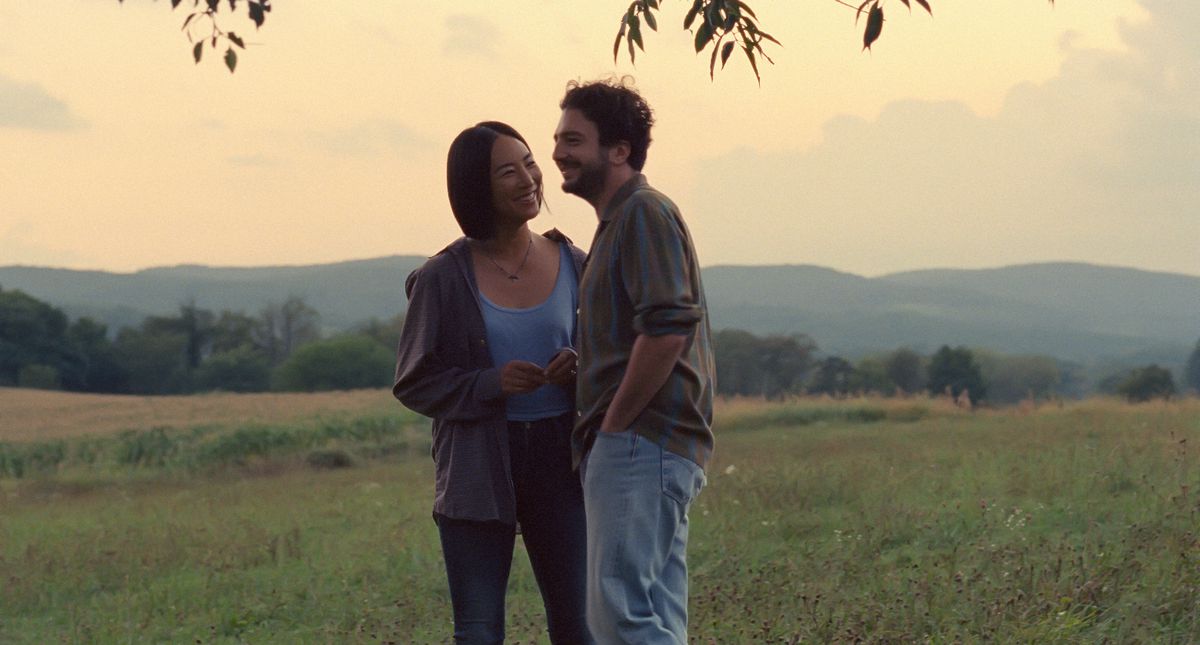 Two actors stand in a field smiling with the sunset behind them. 