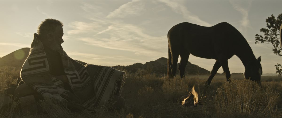 Kidd (Tom Hanks) covered in a blanket lays in a field looking at his horse as the sun sets in News of the World