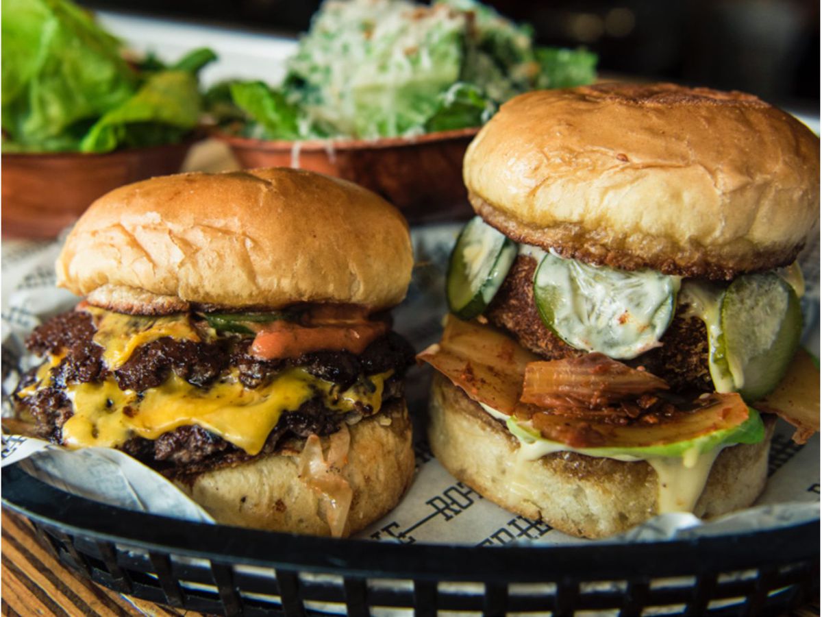 Two high-stacked burgers, one outfitted with melted cheese, the other surrounded by pickles and kimchi.