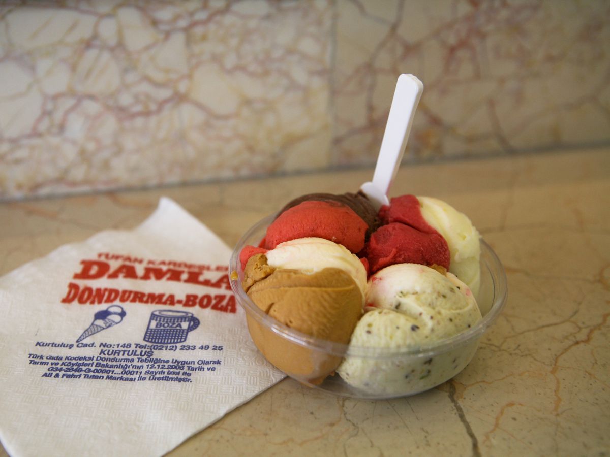 Scoops of colorful ice cream crammed into a clear plastic bowl with a spoon, on a counter with a branded napkin.