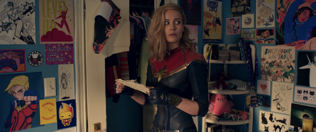 Carol Danvers (Brie Larson) looks confused in Kamala Khan’s bedroom, which is decorated with Carol Danvers memorabilia and fanart in Ms. Marvel.