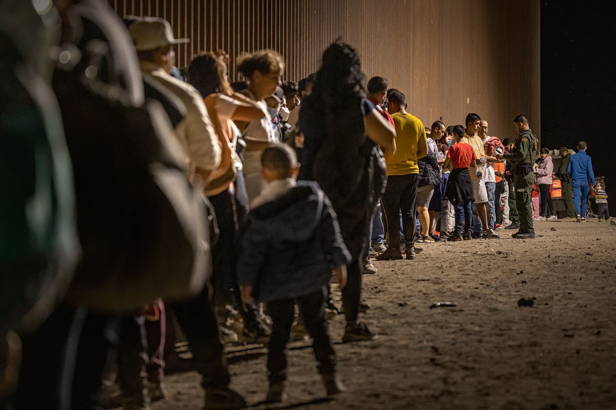 In early morning darkness, a long line of people, several children among them, wait by a tall brown wall outdoors, while uniformed Border Patrol officers talk to those in the front.  