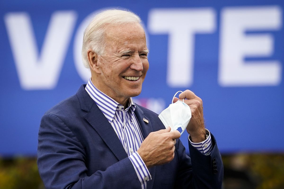 Biden, in a navy suit jacket, a dress shirt with bold vertical blue and white stripes, and no tie, smiles as he removes a white mask. He stands in the sunlight in front of a massive blue sign that reads, in bold white font, “VOTE.”