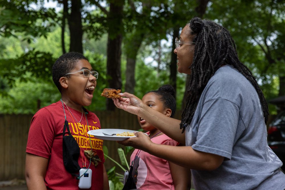 Smiling Black woman feeds a piece of fried chicken to a child standing with their mouth open; another little girl watches.