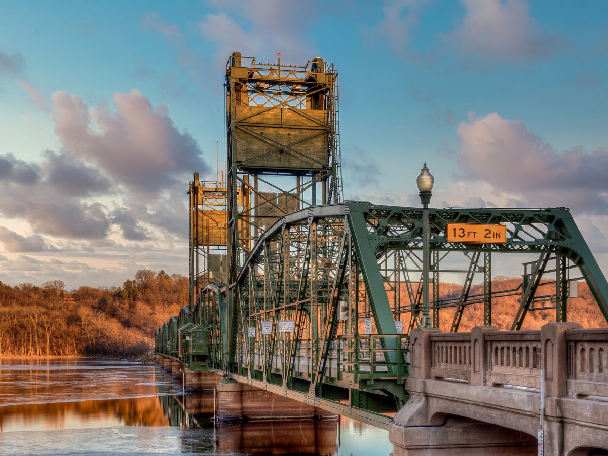 An old-fashioned lift bridge stretches across an expanse of peaceful water at sunset; trees are visible in the background. 